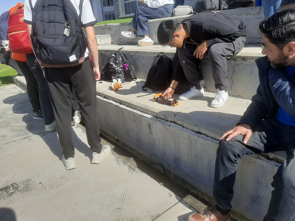 Estudiantes universitarios de la UMAD observan con atención mientras uno de sus compañeros lanza un carrito impulsado por ligas sobre una rampa.